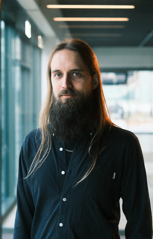 A man with dark beard and long blond hair standing in a concrete and glass building.