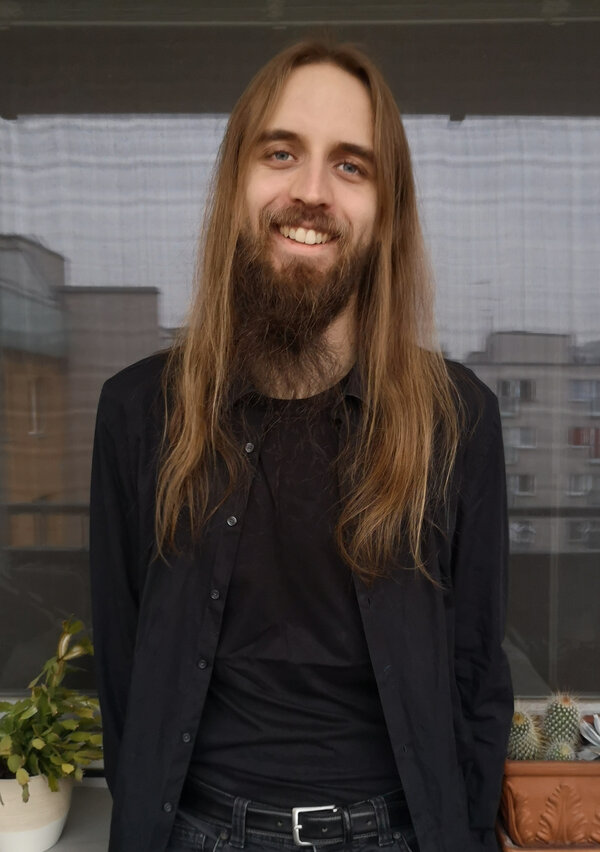 A man with dark beard and long blond hair standing in front of reflections in a window